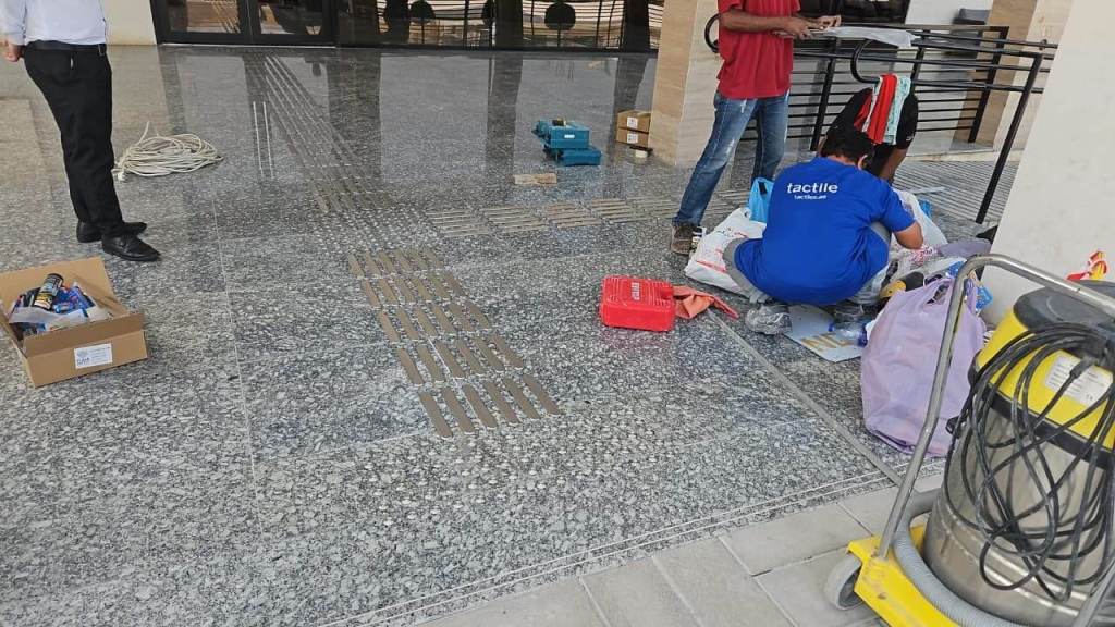 A group of workers on a polished stone floor, one sitting and sorting items, while another stands nearby. Tools and cleaning equipment are visible, alongside boxes and materials scattered around.