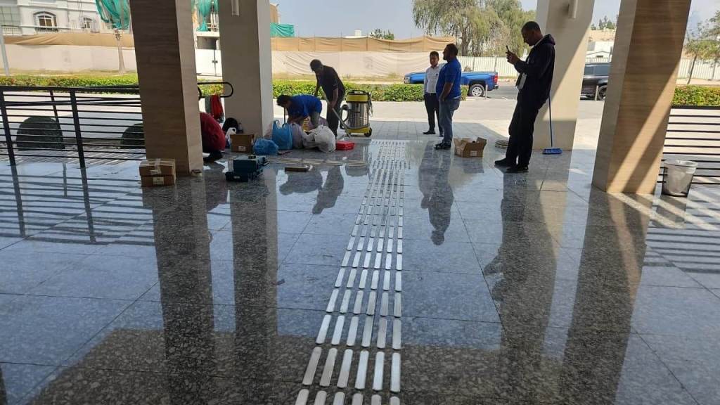 An interior view of a lobby with polished marble flooring. Several people are engaged in activities such as setting up equipment and checking their phones. Various tools and materials are scattered on the floor.