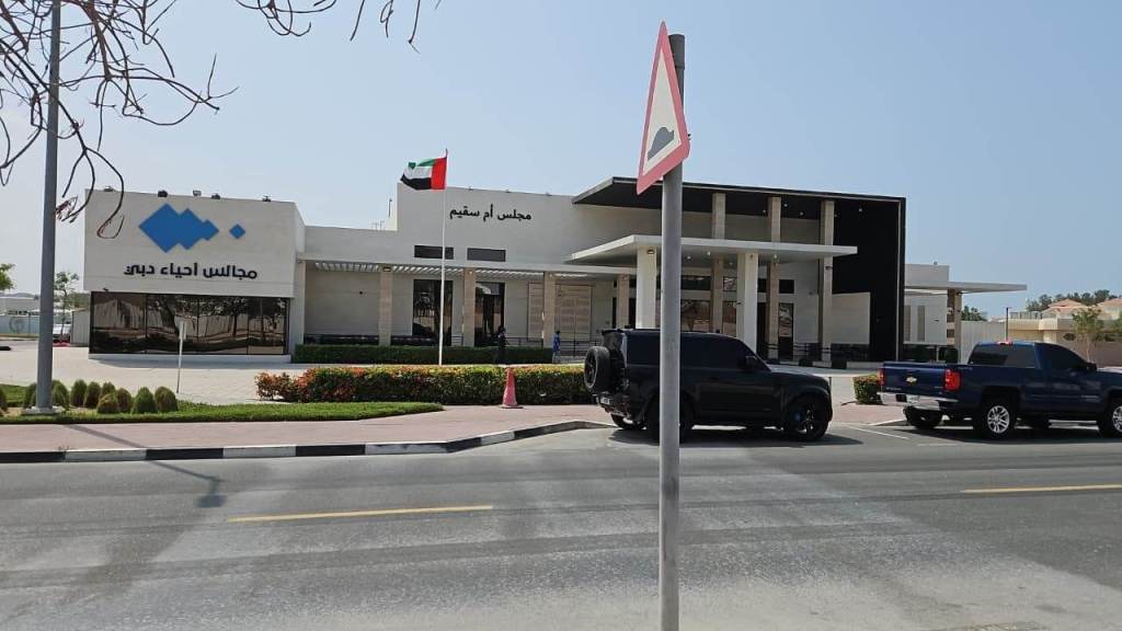 Exterior view of a modern building with a UAE flag, featuring a sign in Arabic that reads 'مجالس أم سقيم', located along a road with parked cars in front.