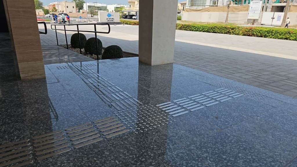 A concrete walkway with textured surface features, including tactile paving and a ramp leading to an entrance, surrounded by greenery and buildings in the background.