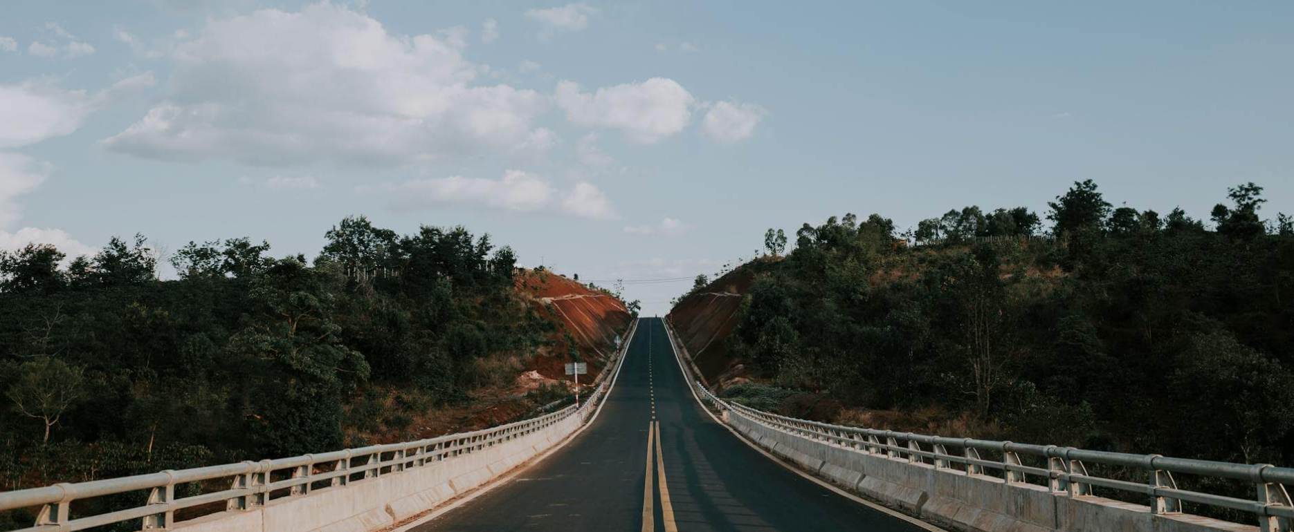 gray concrete road between green trees under white clouds and blue sky