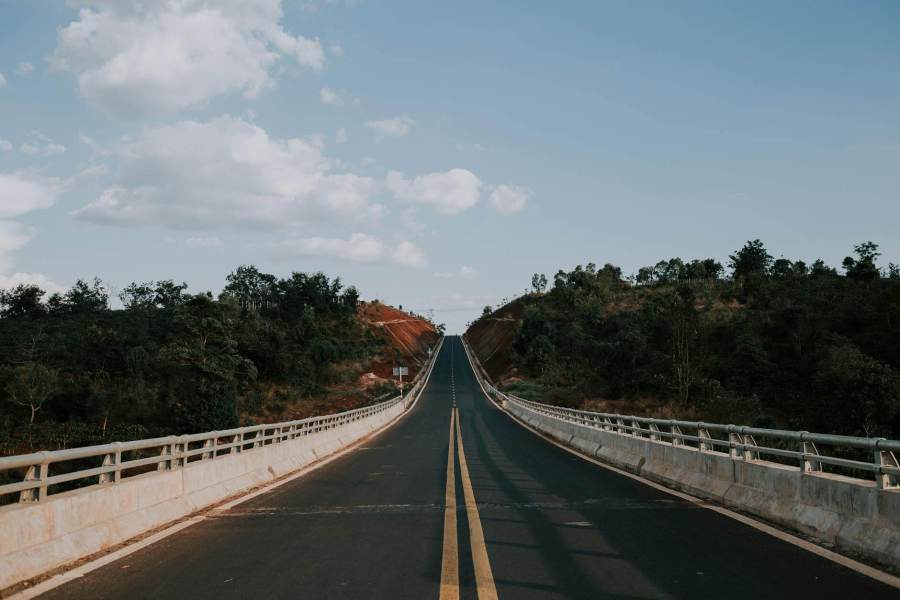 gray concrete road between green trees under white clouds and blue sky