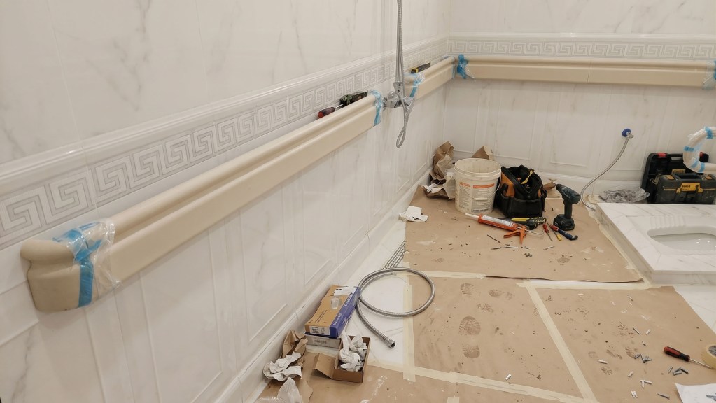 A partially renovated bathroom with marble-patterned tiles, featuring a beige trim along the walls. Tools, construction materials, and debris are scattered on the floor.